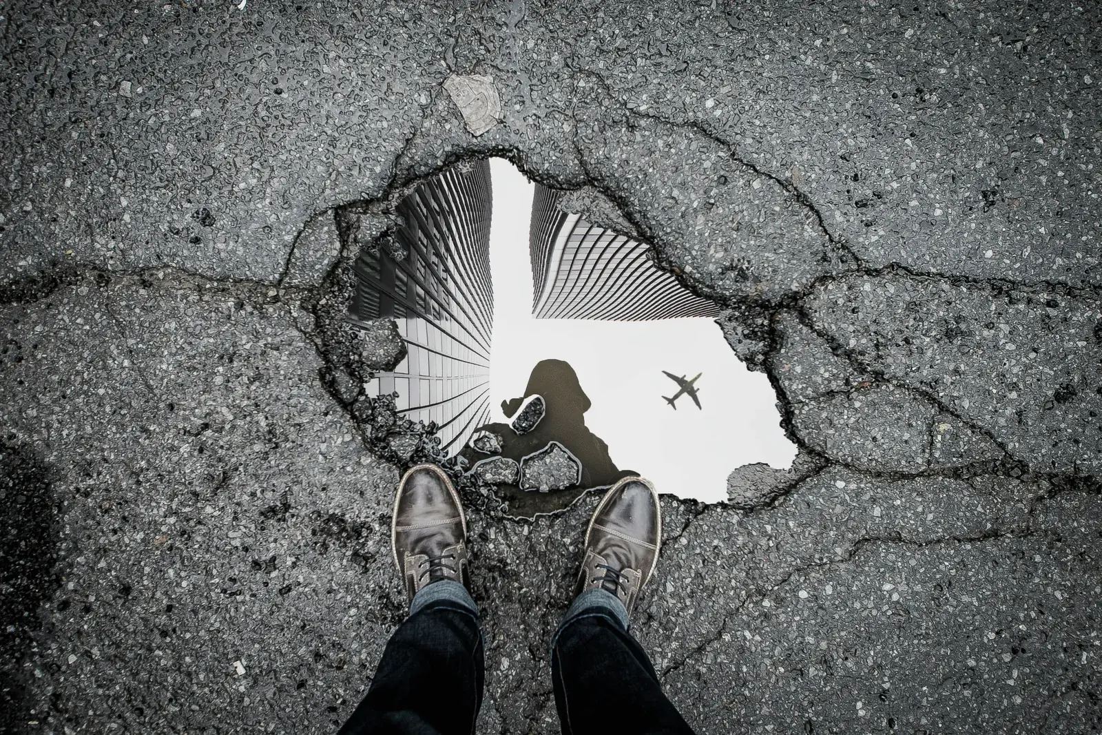 Pessoa observa o reflexo de edifícios e de um avião em uma poça sobre o asfalto.