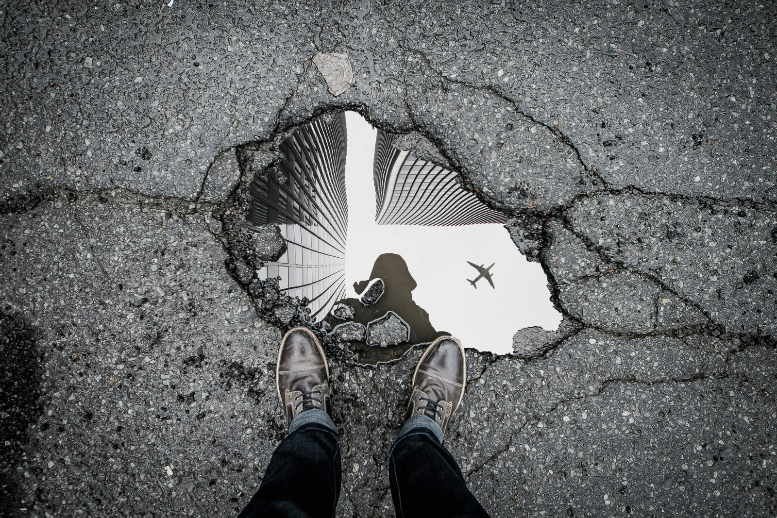 Pessoa observa o reflexo de edifícios e de um avião em uma poça sobre o asfalto.
